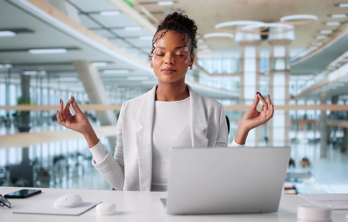 stress-relief | Professional woman practicing mindfulness meditation at office desk with laptop, demonstrating stress management techniques for paralegals