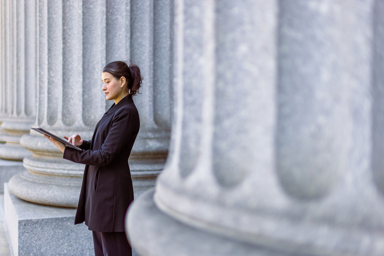 | Professional paralegal reviewing legal documents on tablet outside New York City courthouse with classical columns